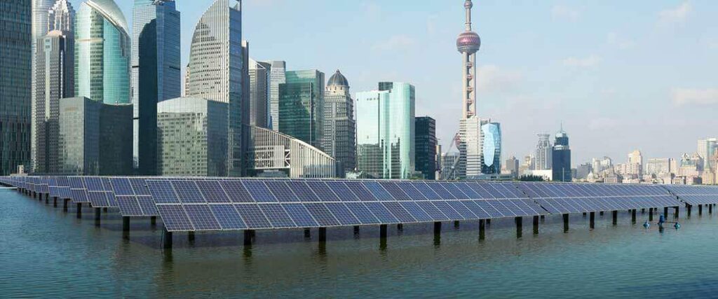 Rows of solar panels mounted on posts in the sea in front of a modern metropolis with numerous skyscrapers.