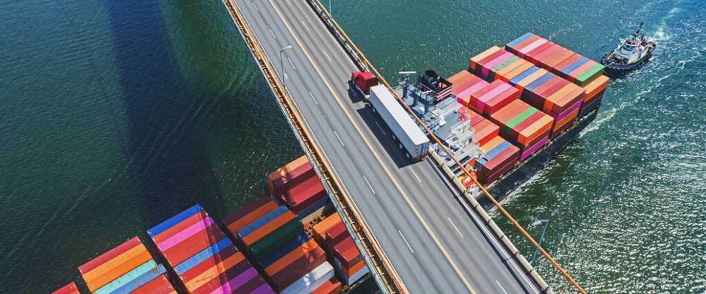 An overhead view of a loaded semi-truck crossing a bridge, under which is a cargo container ship.
