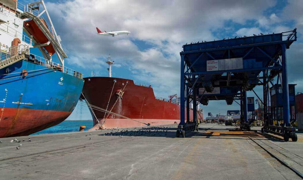 A passenger jet flying over cargo ships and loading platforms at port.