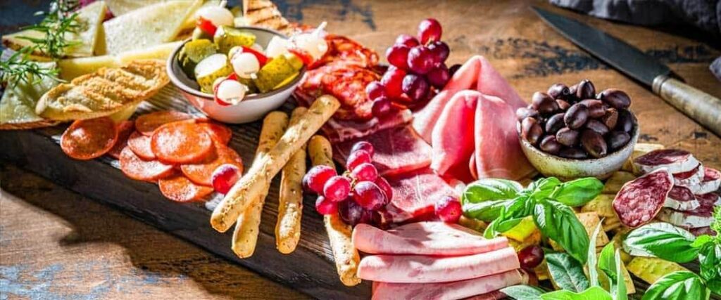 A charcuterie board including pepperoni, ham, salami, breadsticks, olives, pickles, and more.