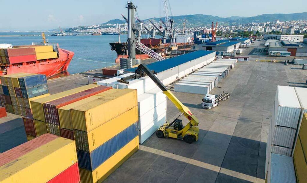 Several shipping containers stacked up at port with a crane removing one to place on a short-range drayage truck.