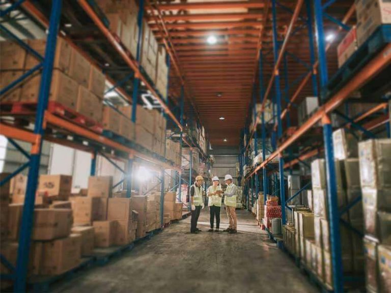 An interior view of a bonded warehouse with two employees in the background and shelves with containerized goods extending to the foreground, reinforcing the article's subject: bonded warehouse vs ftz