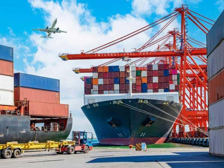 The image shows a container ship docked at port and being loaded with freight.